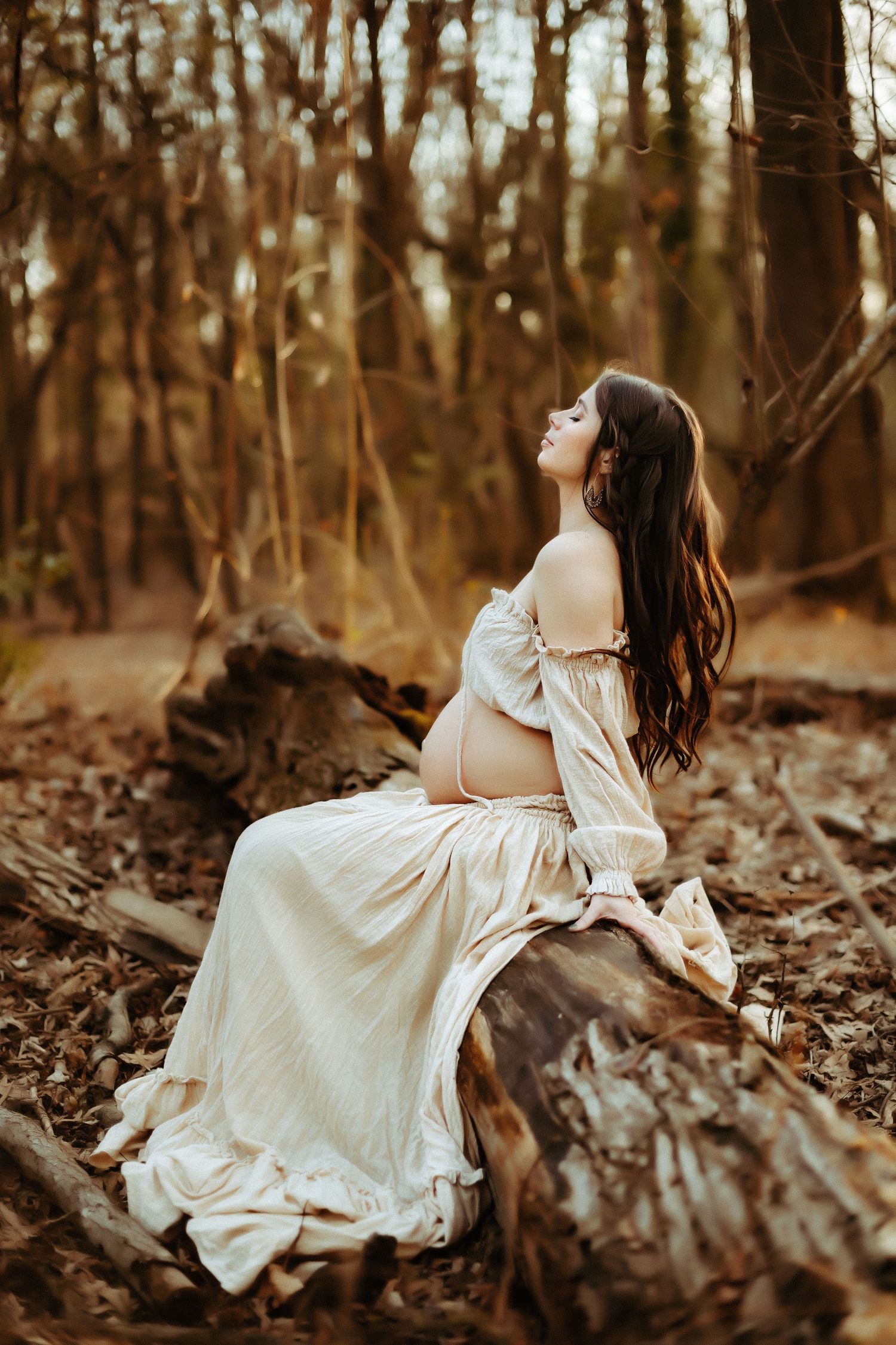 Artistic maternity photo in autumn forest with flowing cream dress sitting on fallen log in golden light.