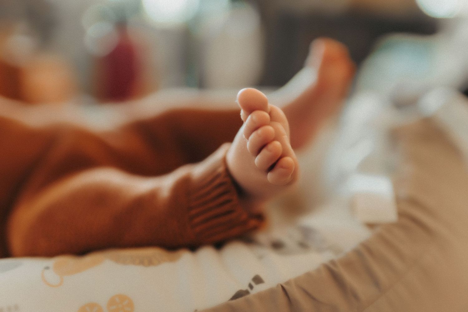Close-up of tiny baby foot with soft brown knit sock against white bedding in warm lighting.