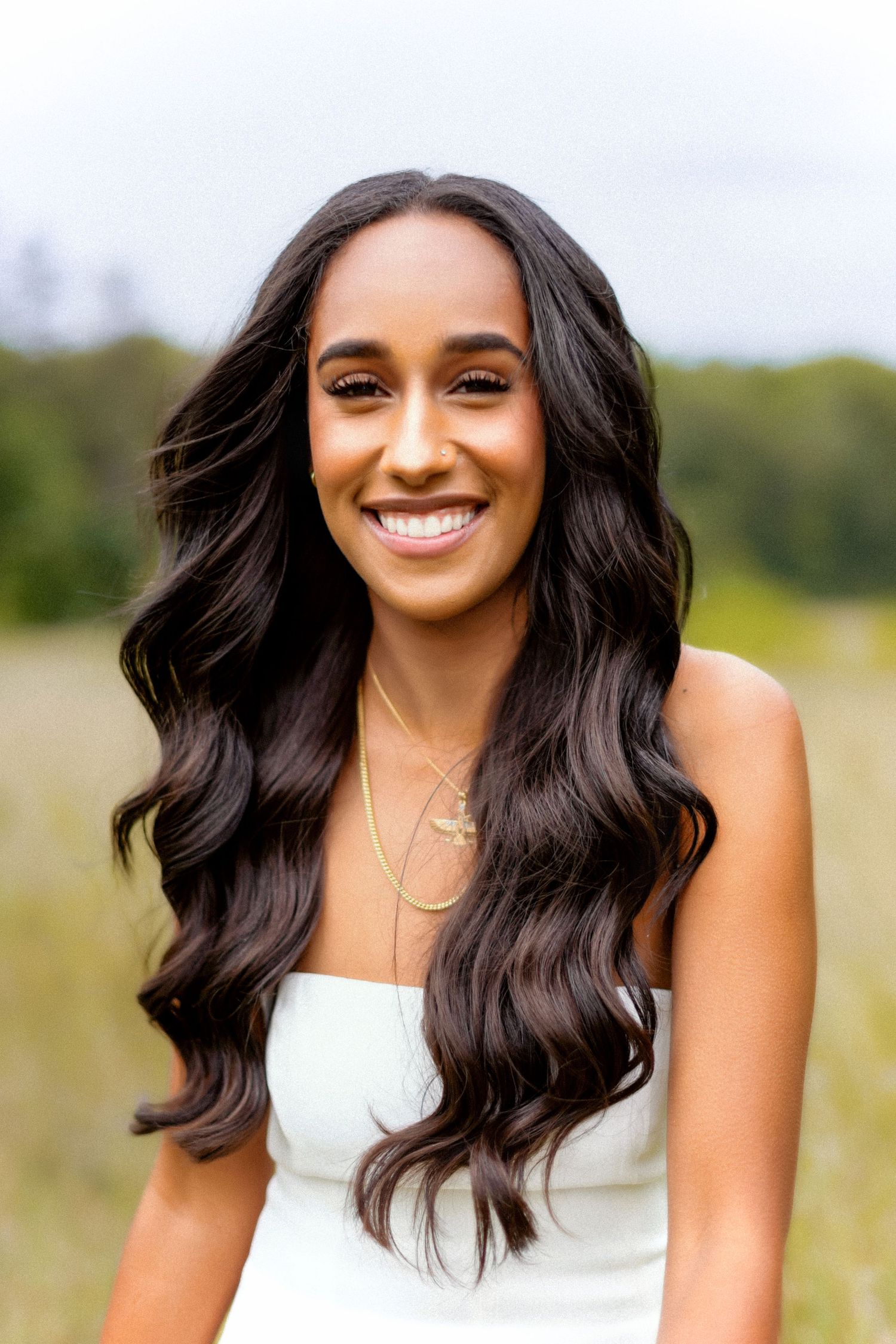 Beautiful outdoor portrait with long wavy dark hair wearing a white dress and delicate necklace in a natural setting.