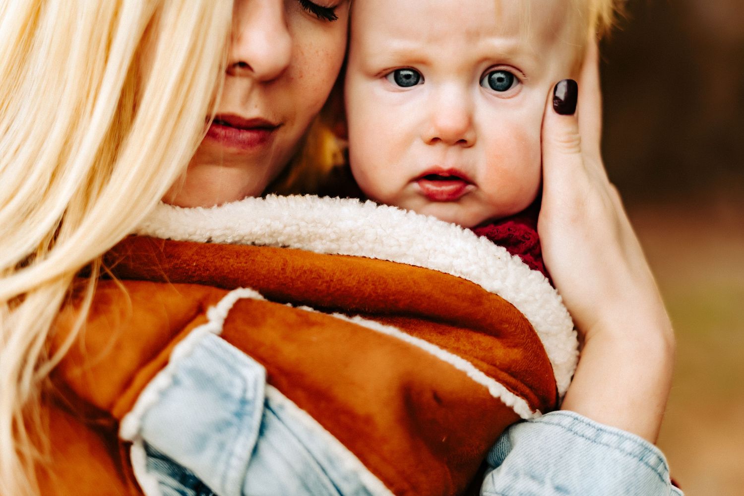 A caring adult holds a small child wearing a brown furry jacket while outdoors on a fall day.