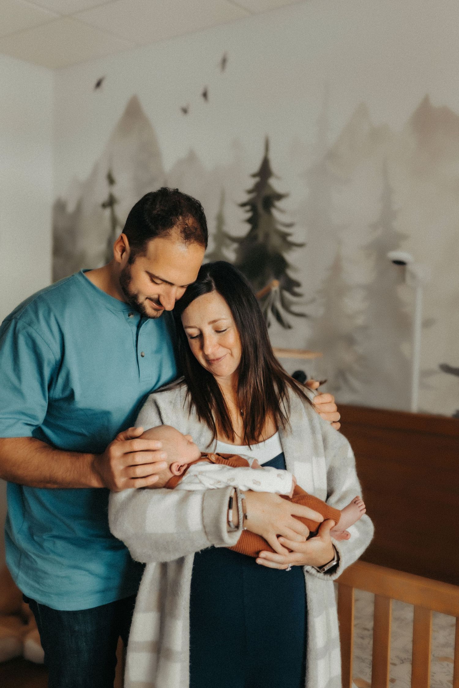 A couple shares an intimate moment with their newborn baby in front of a wall decorated with painted forest scenery.