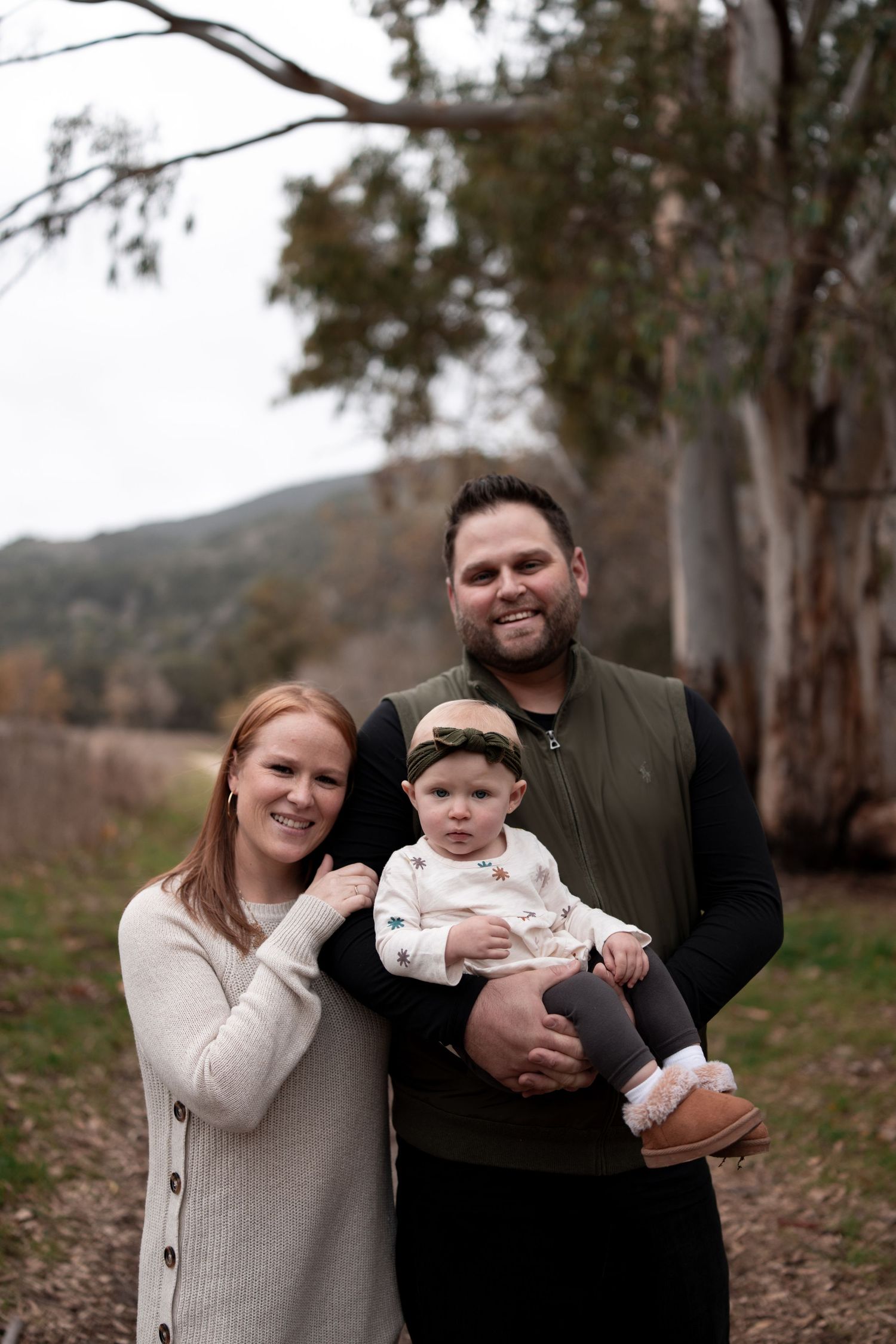 A sequence of four outdoor family photos shows a couple and infant against a backdrop of eucalyptus trees on a cloudy day.