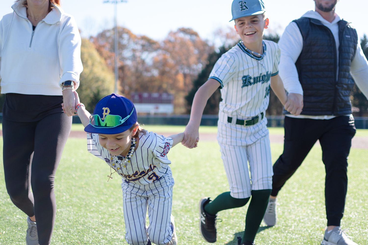 Youth baseball players wearing pinstriped uniforms run together on a grass field during a sunny fall day.