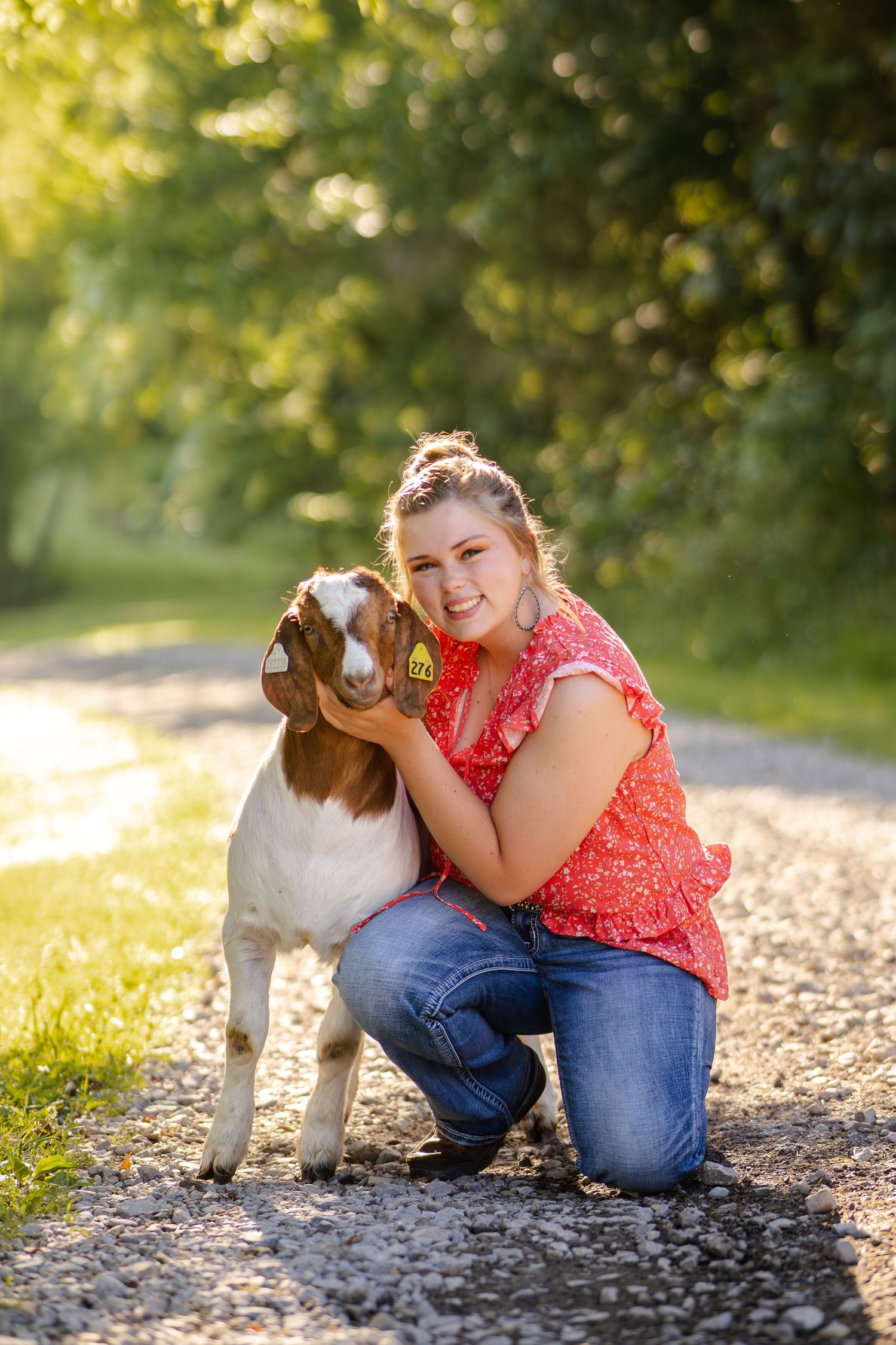Smiling young woman in a red floral top kneeling on a gravel path hugging her market goat.