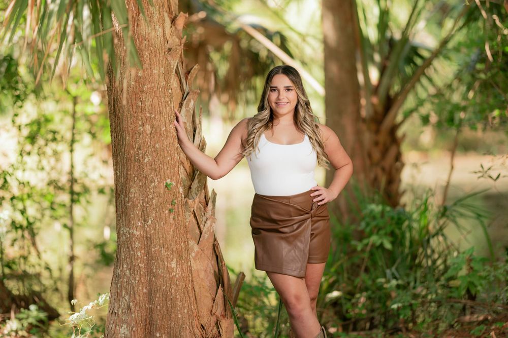 Woman in white tank top and brown leather skirt poses by tree trunk in lush garden setting.
