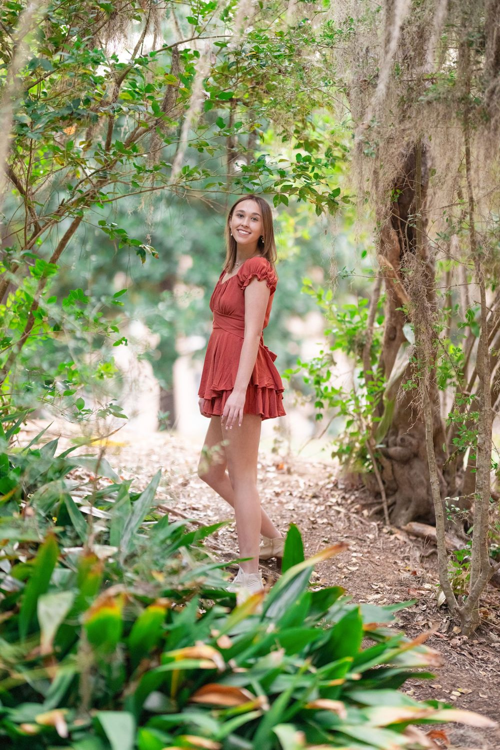 A person in a red ruffled dress poses along a garden path surrounded by lush greenery and trees.