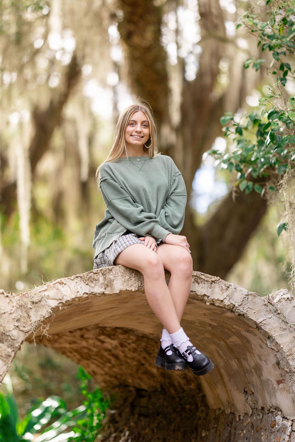 Someone wearing a green sweatshirt and shorts sits casually on a curved tree branch in a sunlit wooded setting.