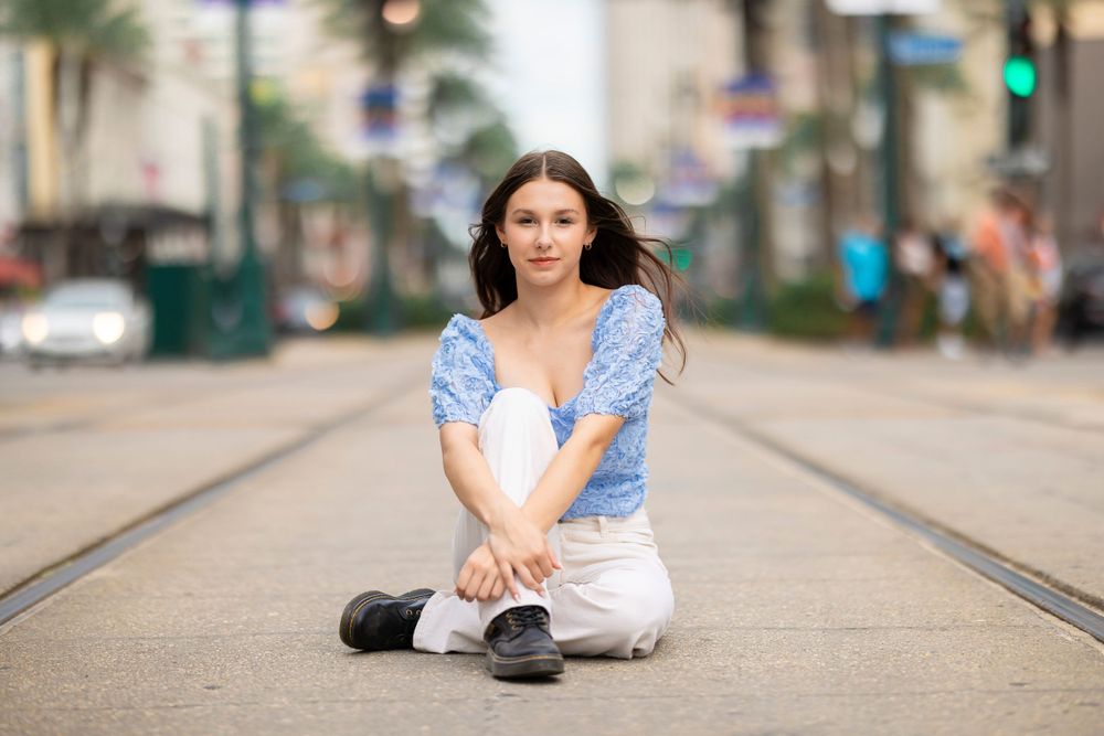 Person in blue puff sleeve top and white pants sits on train tracks in urban setting on sunny day.