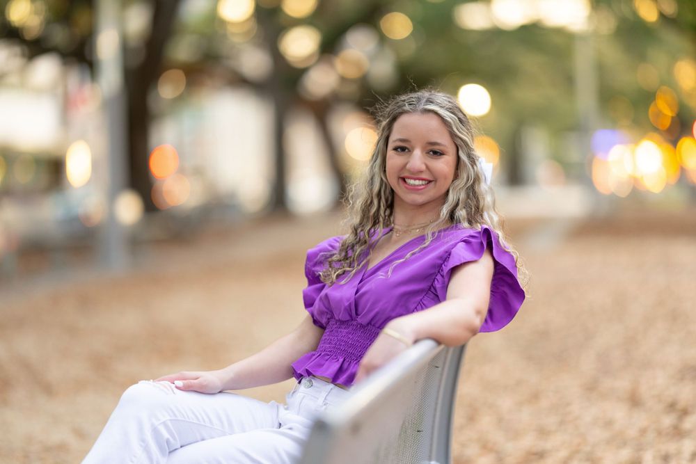 A person in a purple blouse sits on a bench with a blurred bokeh background at golden hour.