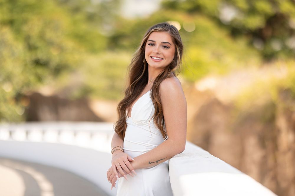 Someone in an elegant white dress poses outdoors along a curved white wall on a sunny day with a blurred nature background.