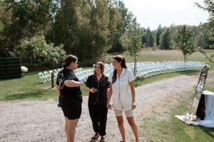 Three people chat casually on a dirt path with white chairs and green trees in the background at an outdoor venue.