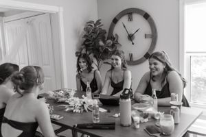 A joyful gathering of people seated around a dining table with a large wall clock and plant decor in the background.