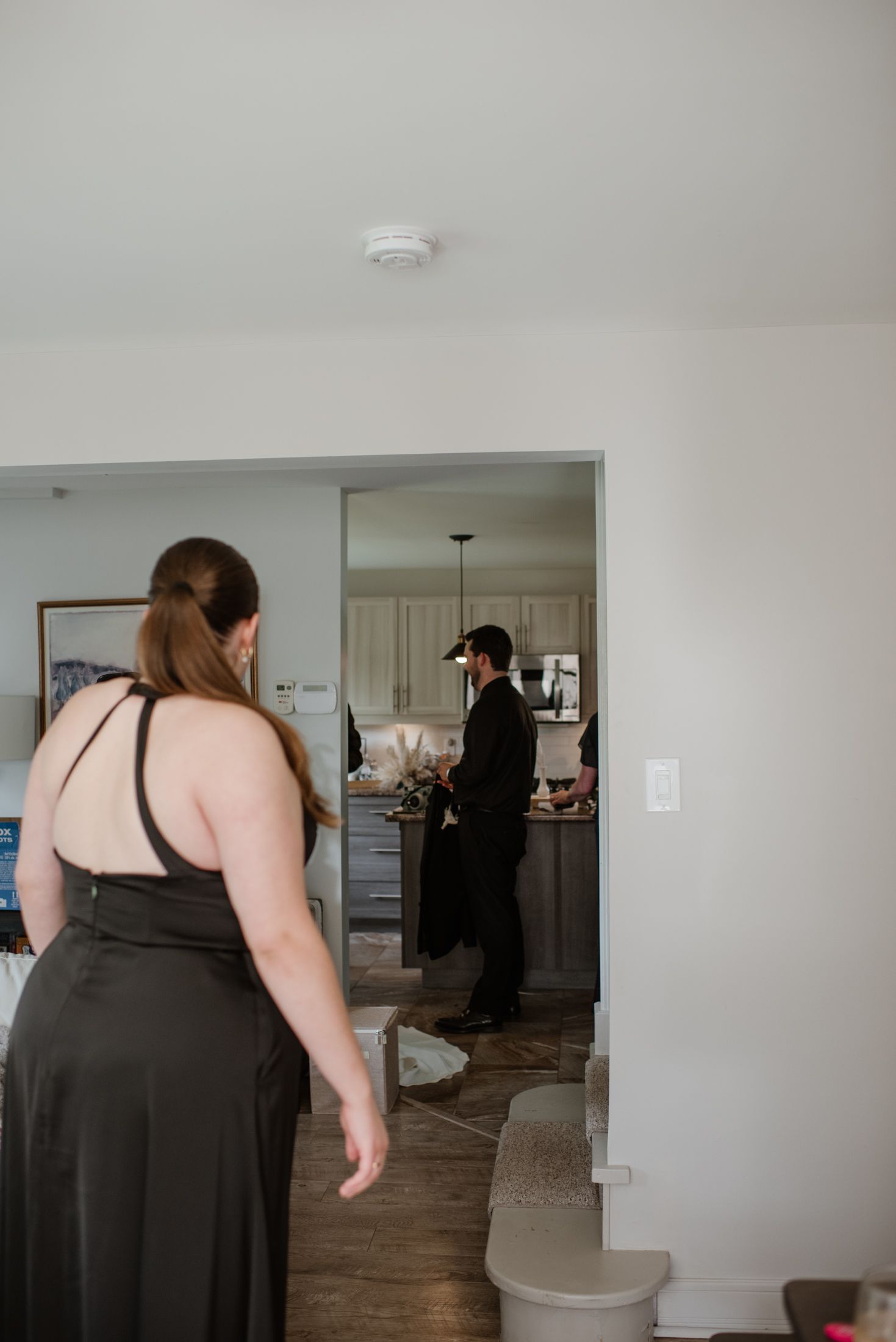 Two people stand in a modern apartment interior with white walls and hardwood floors, one wearing a black dress.