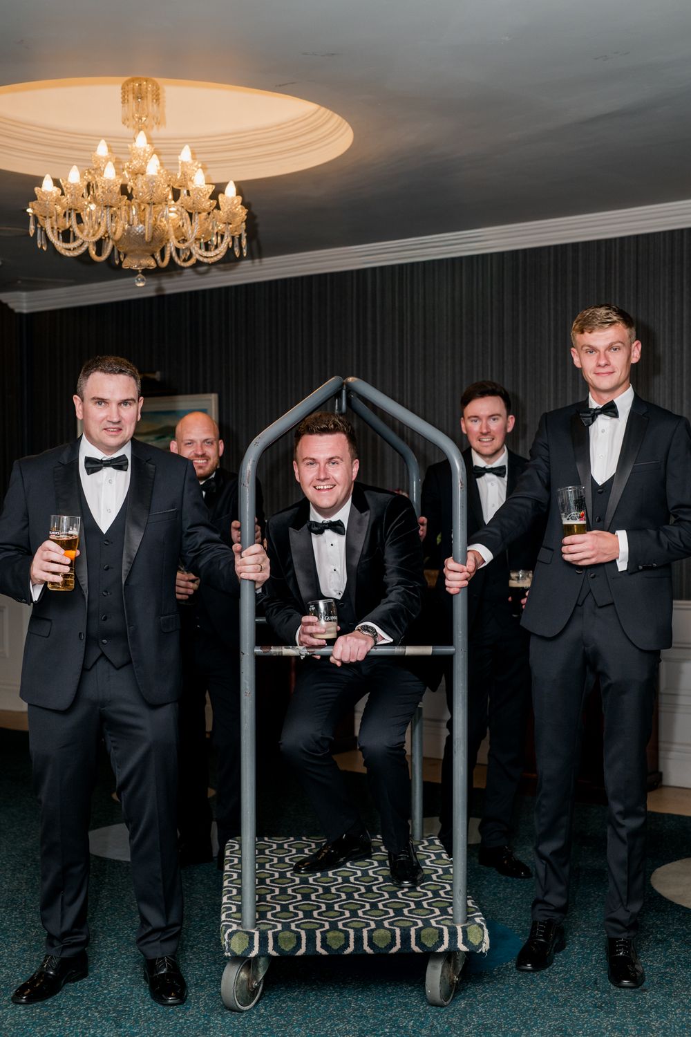 A group in formal black tuxedos and bow ties pose with drinks in an elegant room with chandeliers.