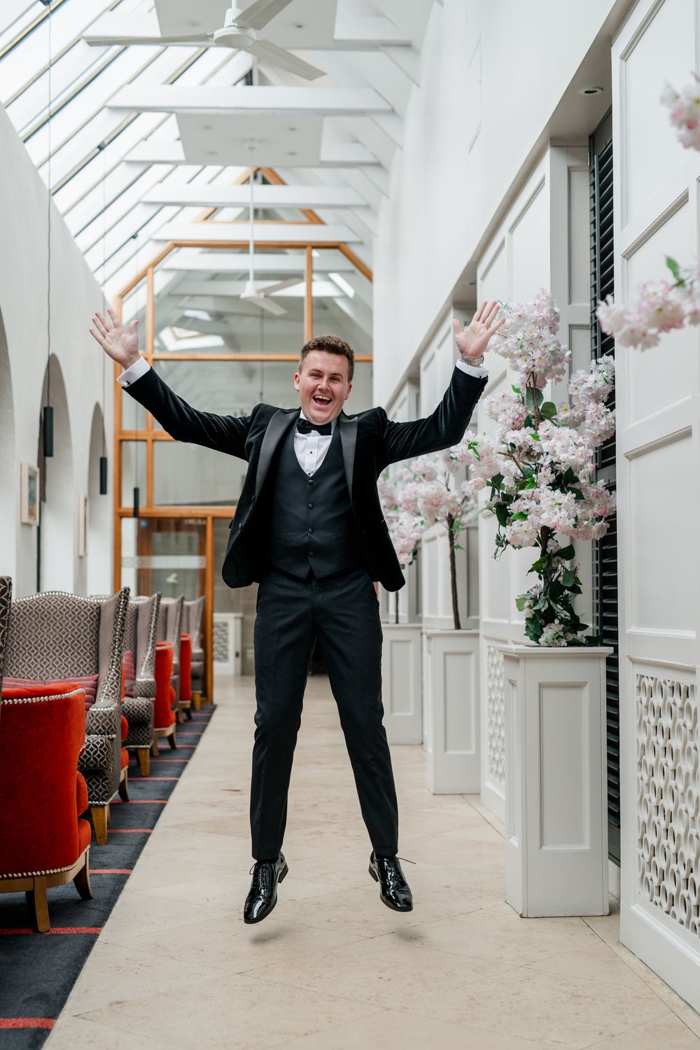 A person in a black tuxedo jumps and poses playfully in a bright white hallway with decorative architectural details.