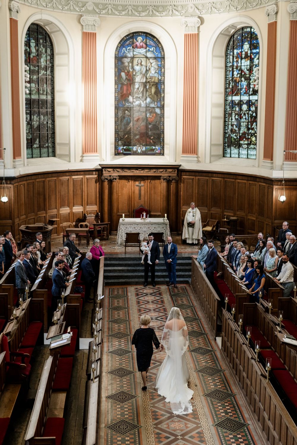A traditional church wedding ceremony with stained glass windows and wooden pews as guests watch a couple walk down the aisle.