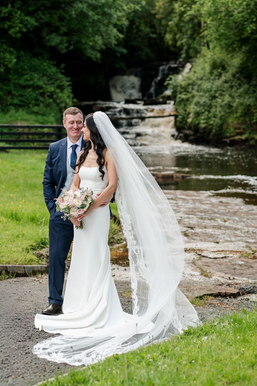 Newlywed couple poses near flowing river and lush greenery for romantic outdoor wedding photos.