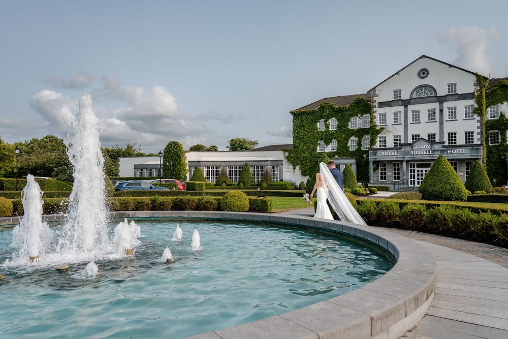 A series of water fountains spray in a curved pool surrounded by manicured gardens and white buildings.