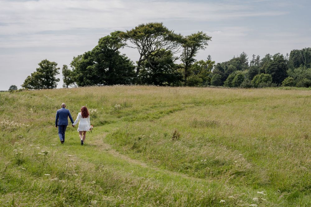 Two people walk together on a grassy hilltop path surrounded by rolling meadows and trees on a sunny day.