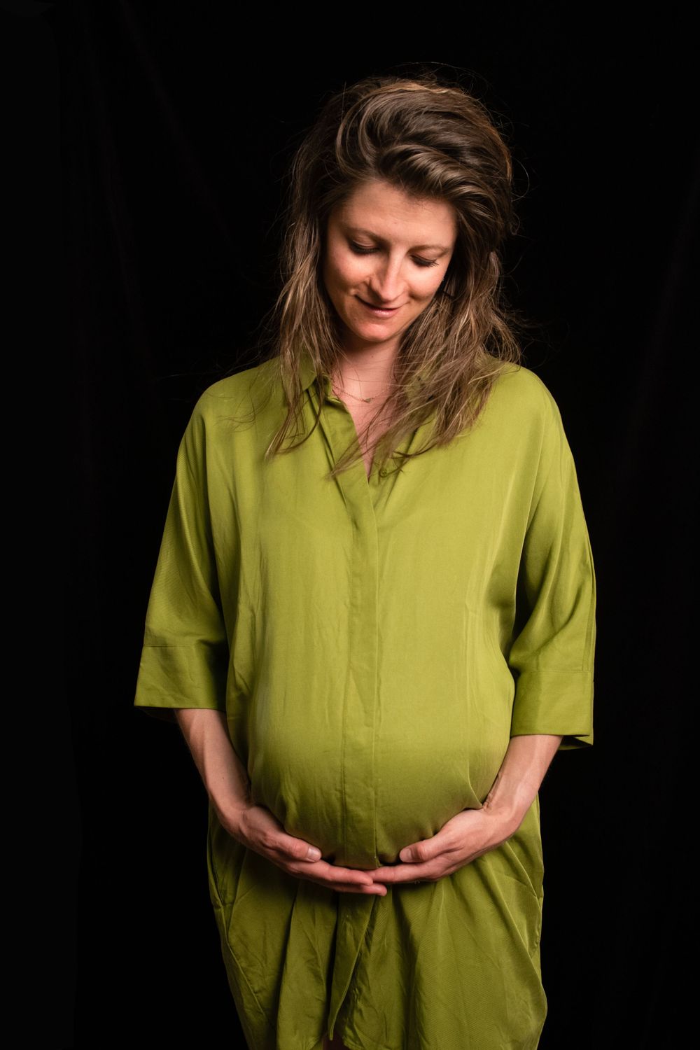 Artistic maternity portrait showing person in lime green dress against black background, cupping her belly.