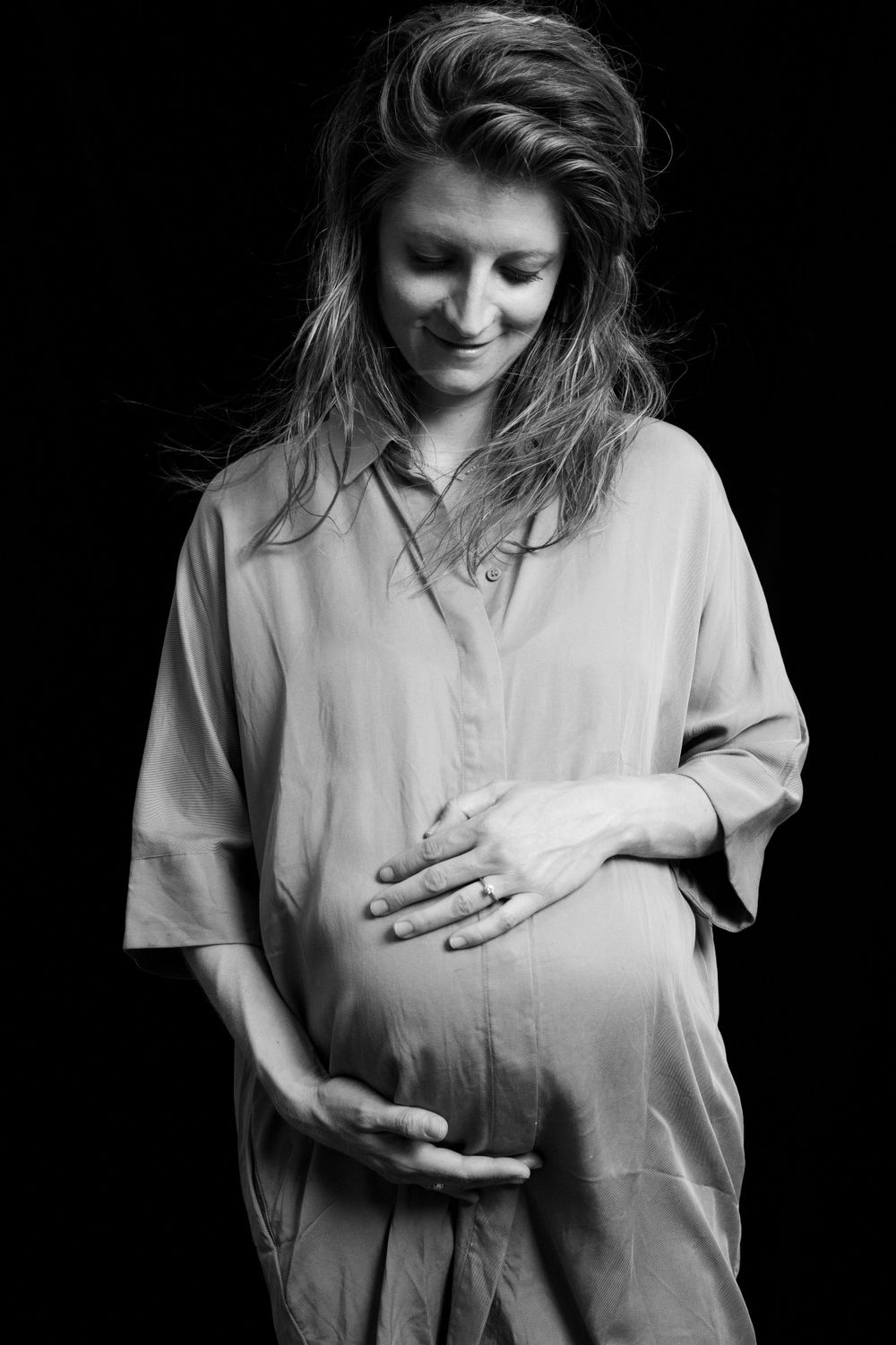 Black and white maternity portrait of person in loose shirt touching belly.