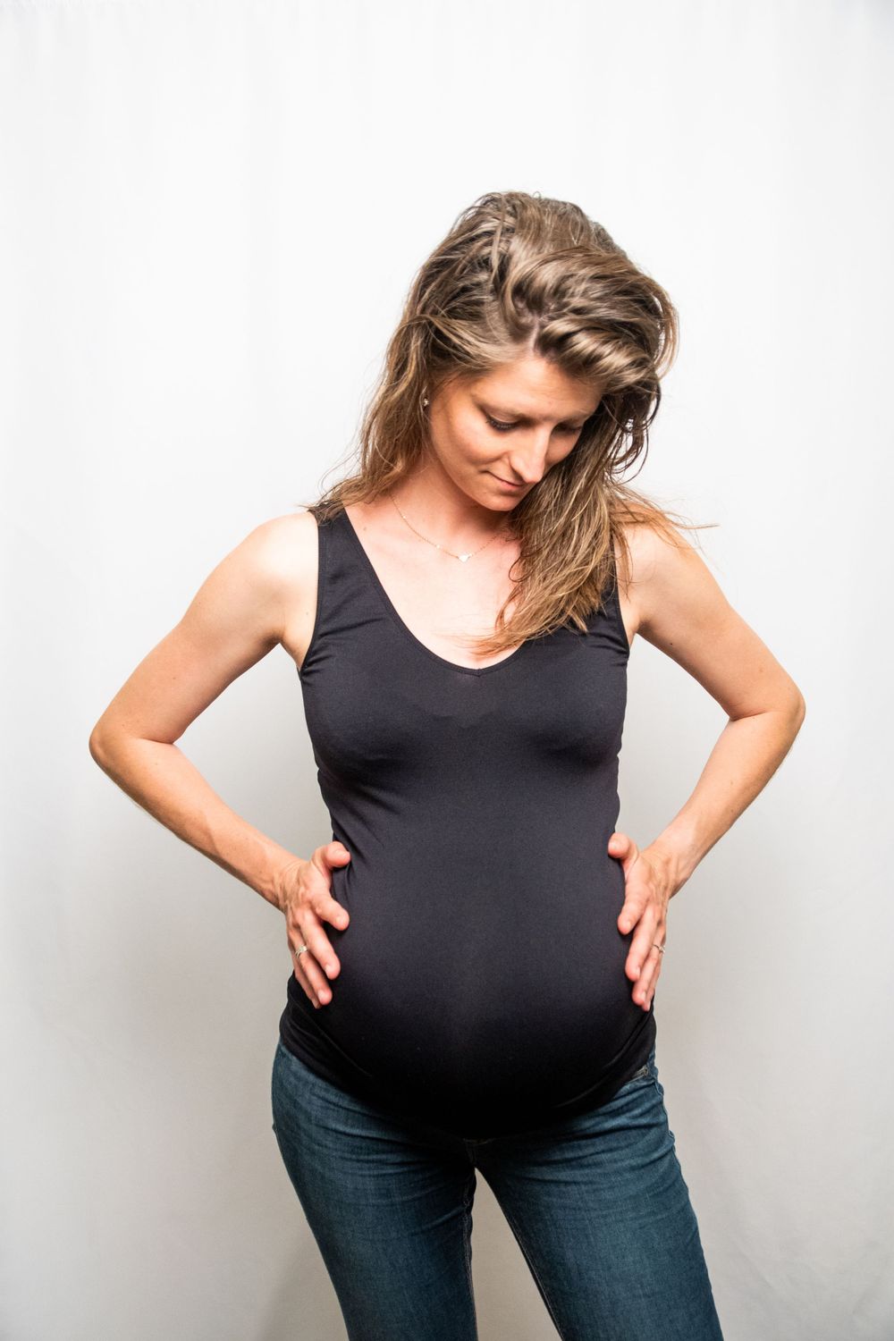 Maternity portrait showing person in black tank top and jeans, looking down at the baby.