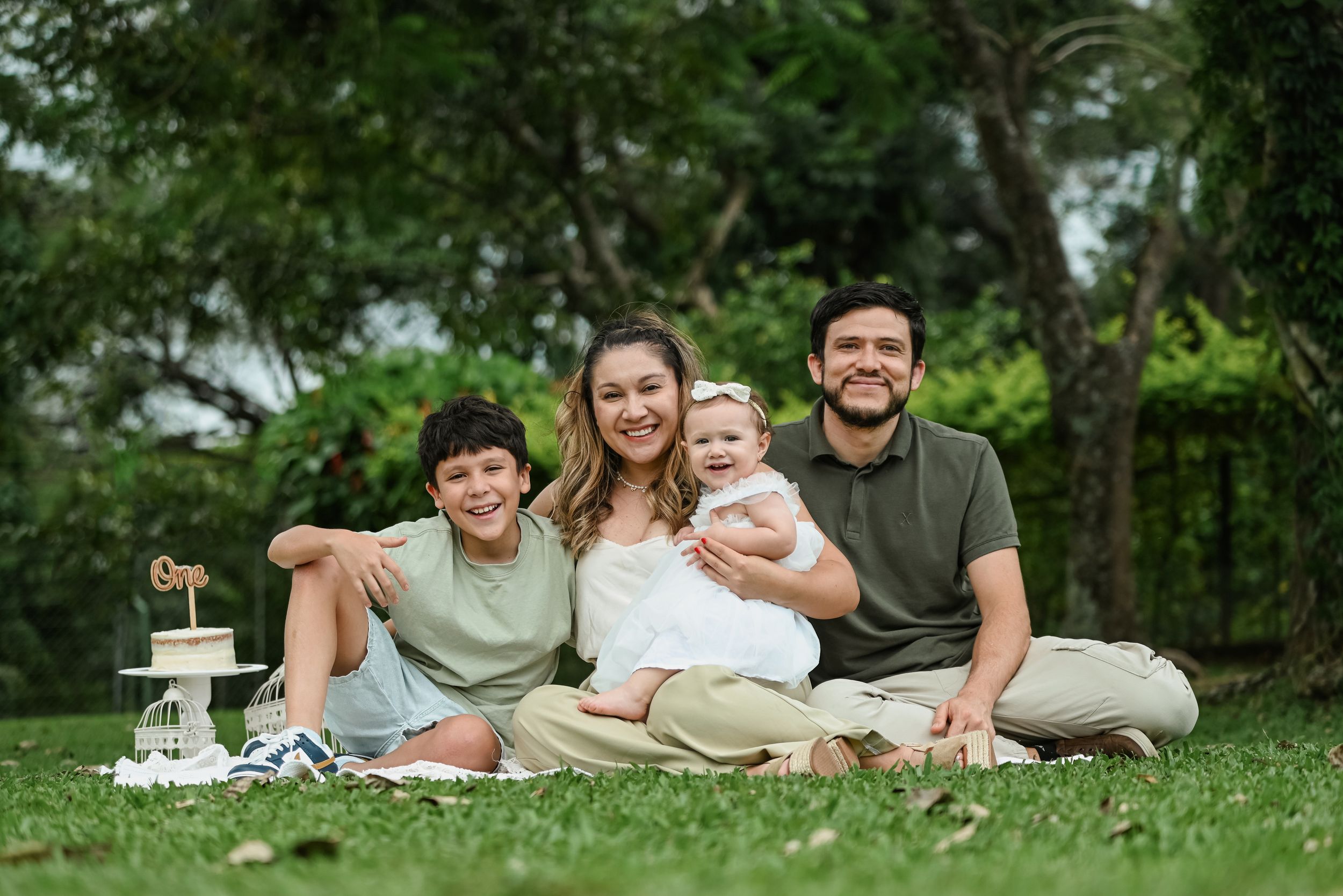 A family sits together on a blanket for a picnic outdoors with trees in the background.