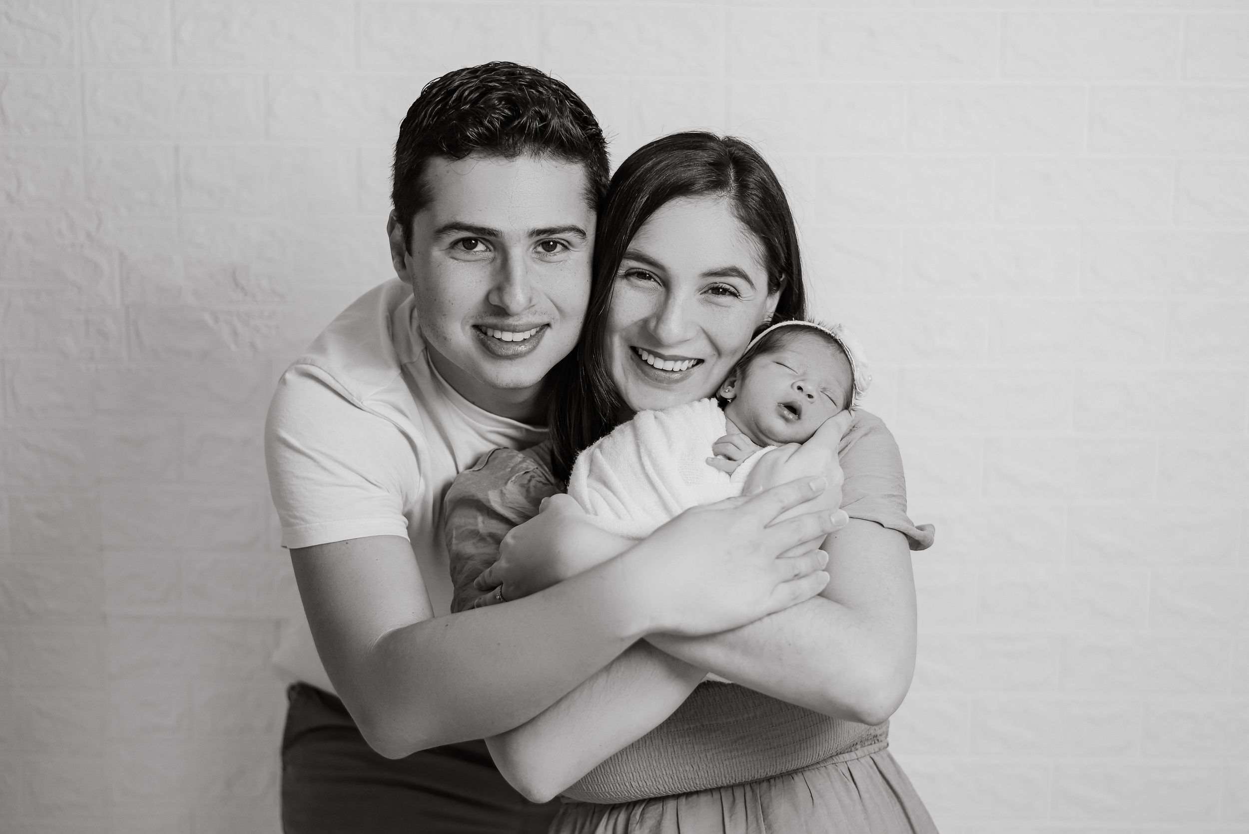 Black and white studio portrait of parents with their newborn baby in a loving embrace against a plain backdrop.