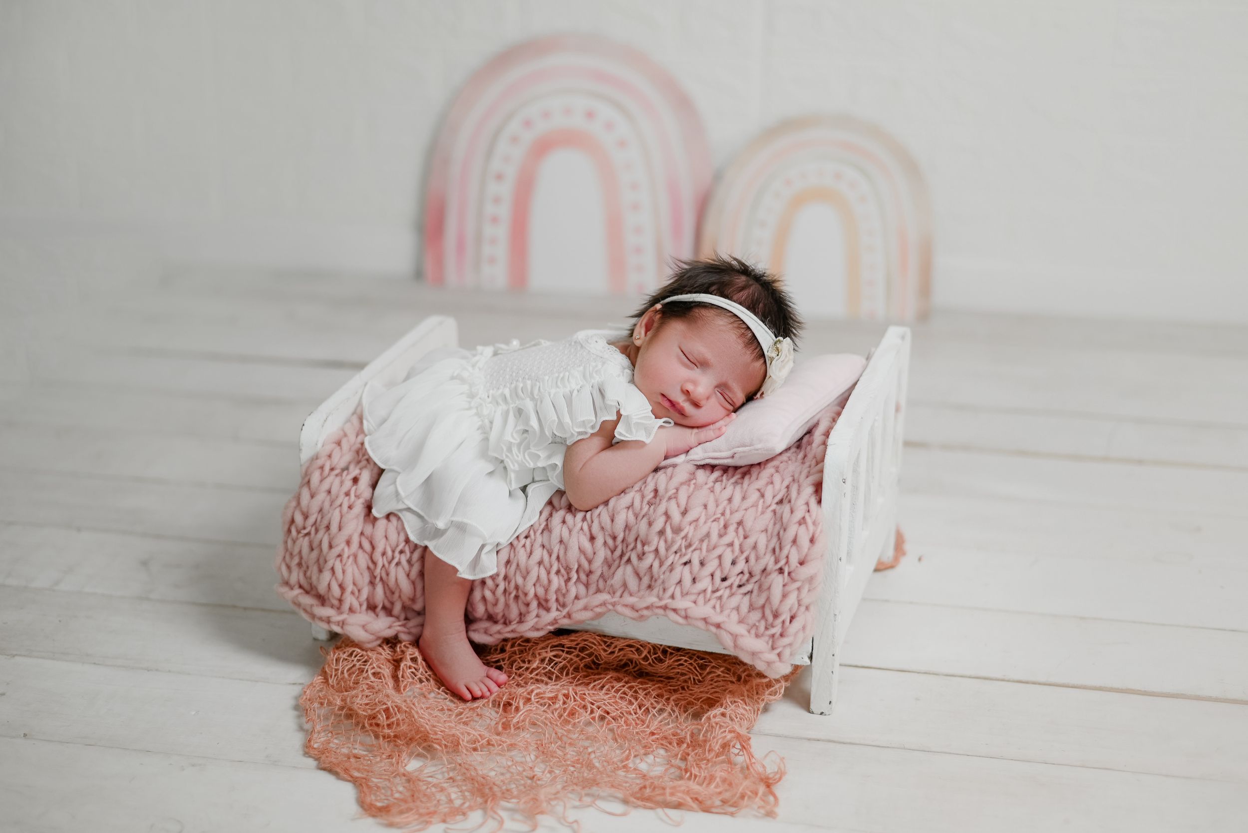 Newborn baby sleeping peacefully on pink bed with white dress and rainbow decorations in background.