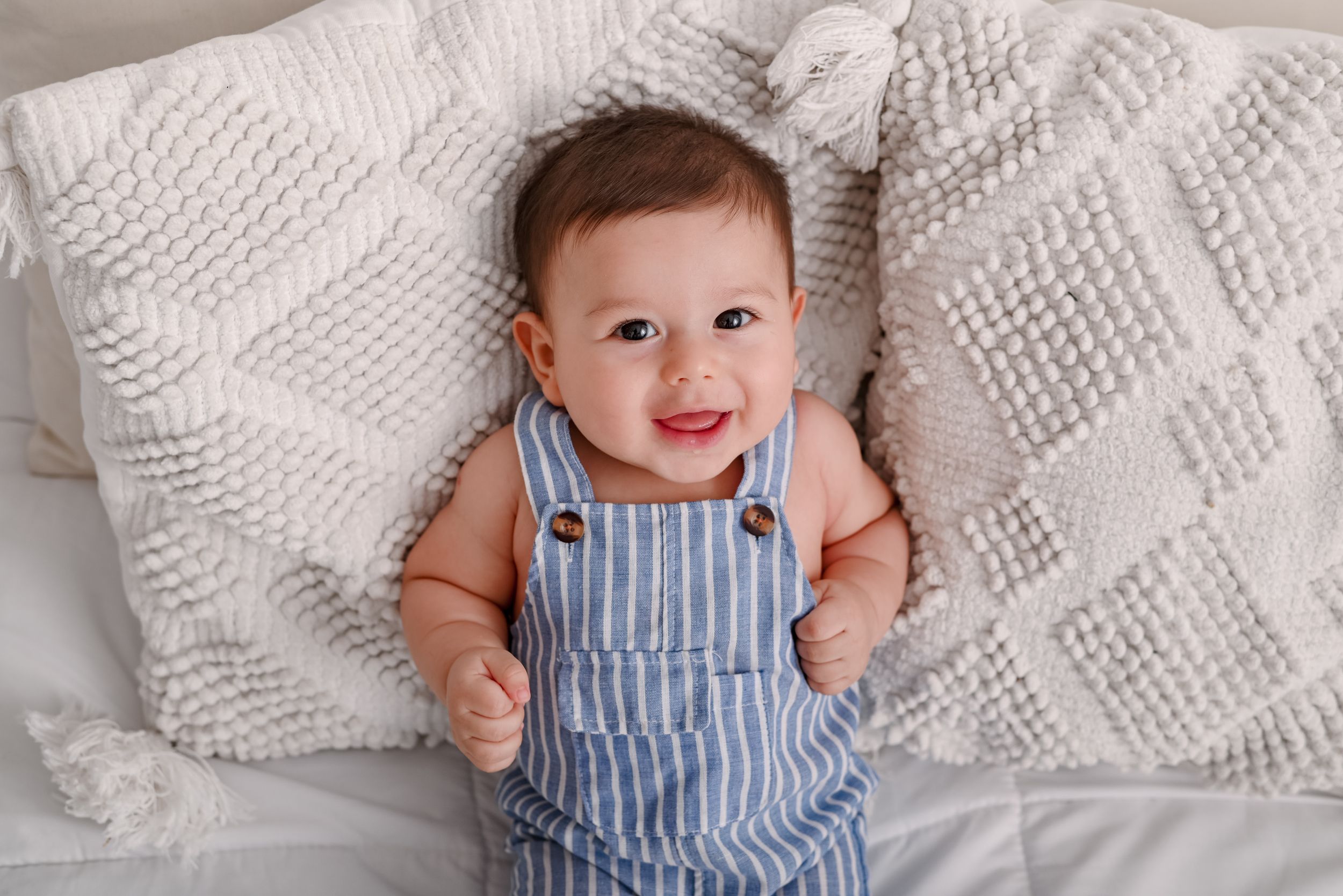 Small baby wearing blue and white striped overalls lies on white knit blanket with a bright happy smile.