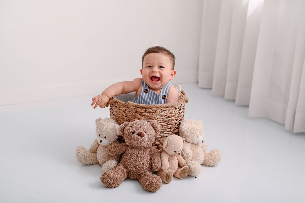 A laughing baby sits in a woven basket surrounded by soft teddy bears on a white floor.