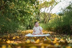 Small child sits on blanket surrounded by fallen autumn leaves and tall grass in a natural outdoor setting.