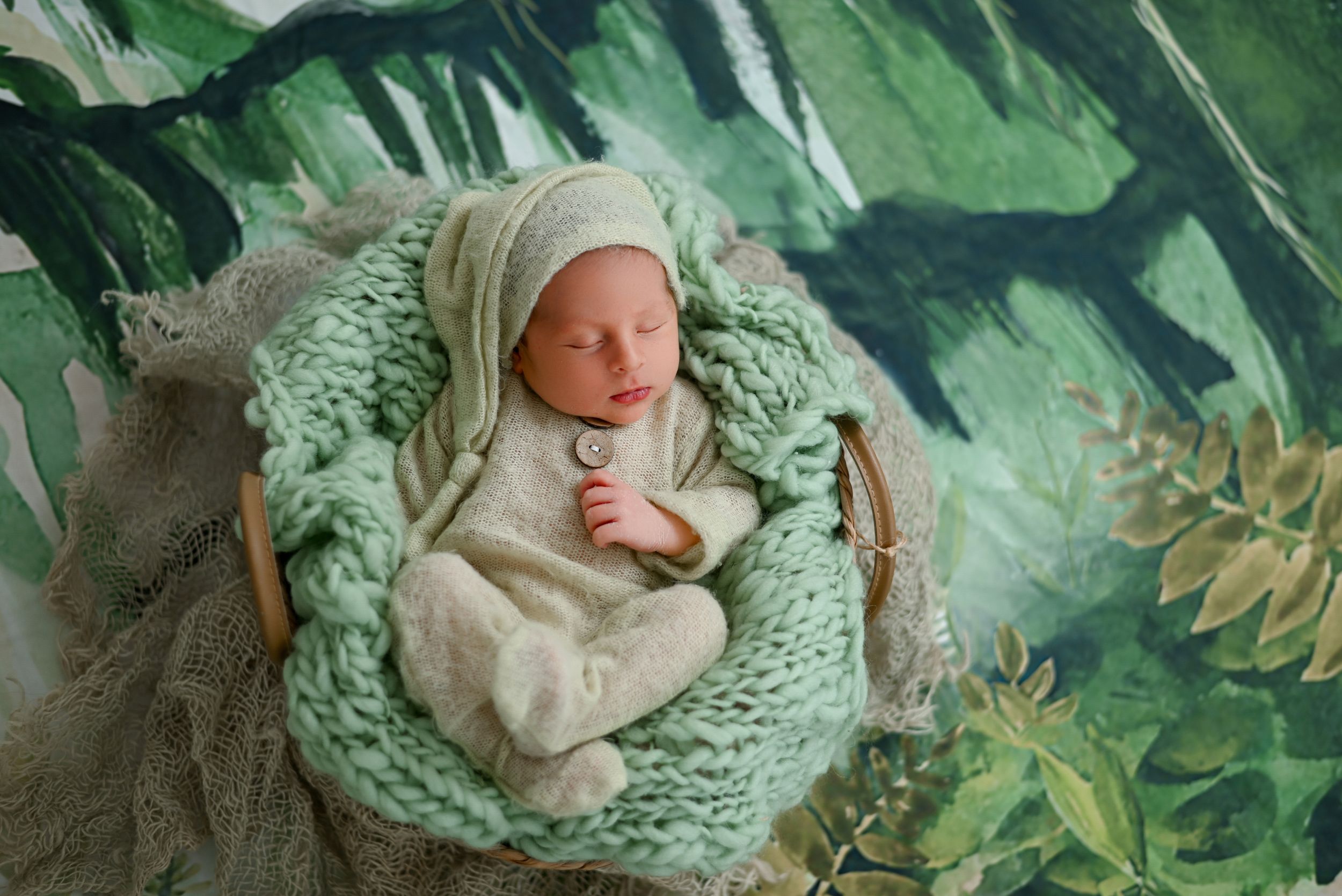 Peacefully sleeping newborn rests in cozy mint green knit blanket surrounded by tropical greenery backdrop.