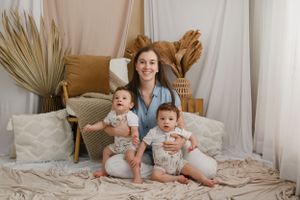 Three people sit together on a cozy bed with beige pillows and dried palm fronds in the background.