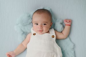 Baby wearing white overalls lies on soft mint green blanket with arm raised.