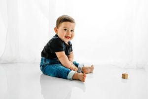 Baby dressed in black t-shirt and blue jeans sits on white floor with wooden toy block.