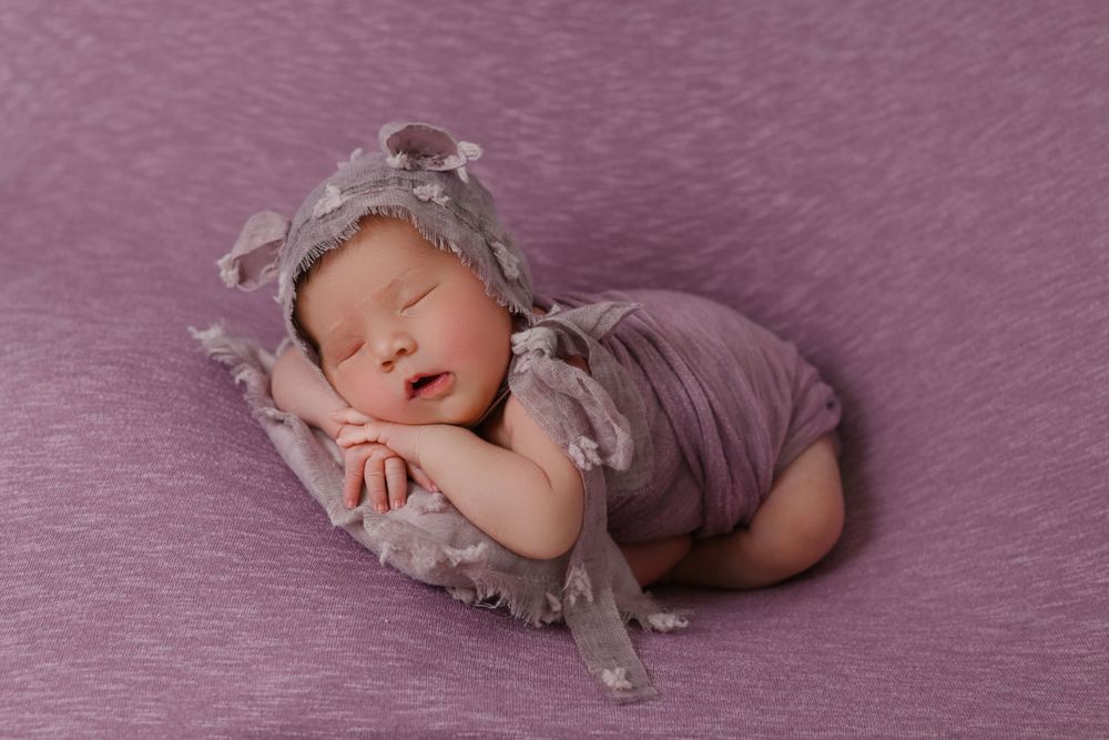 A sleeping newborn in gray attire lies peacefully on a mauve blanket during a professional photo session.