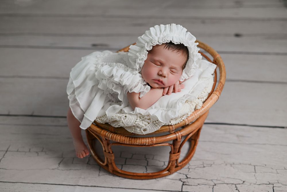A sleeping newborn wearing white lace dress and bonnet rests peacefully in a woven rattan basket on grey wooden floor.
