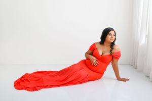 Woman in flowing red off-shoulder gown poses elegantly on white floor near window.