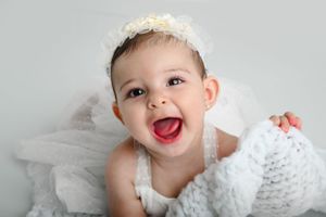 A baby wearing white lace and tulle shows a joyful expression while laying on a gray background.
