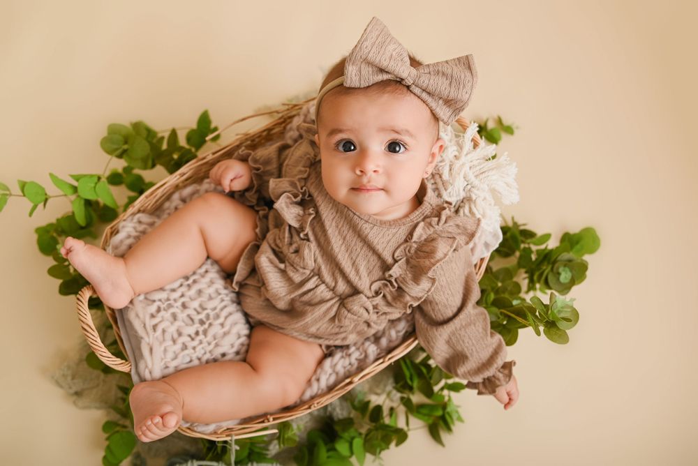 A small infant wearing beige ruffled clothing and bow lies on greenery in a rustic wooden basket for a portrait session.