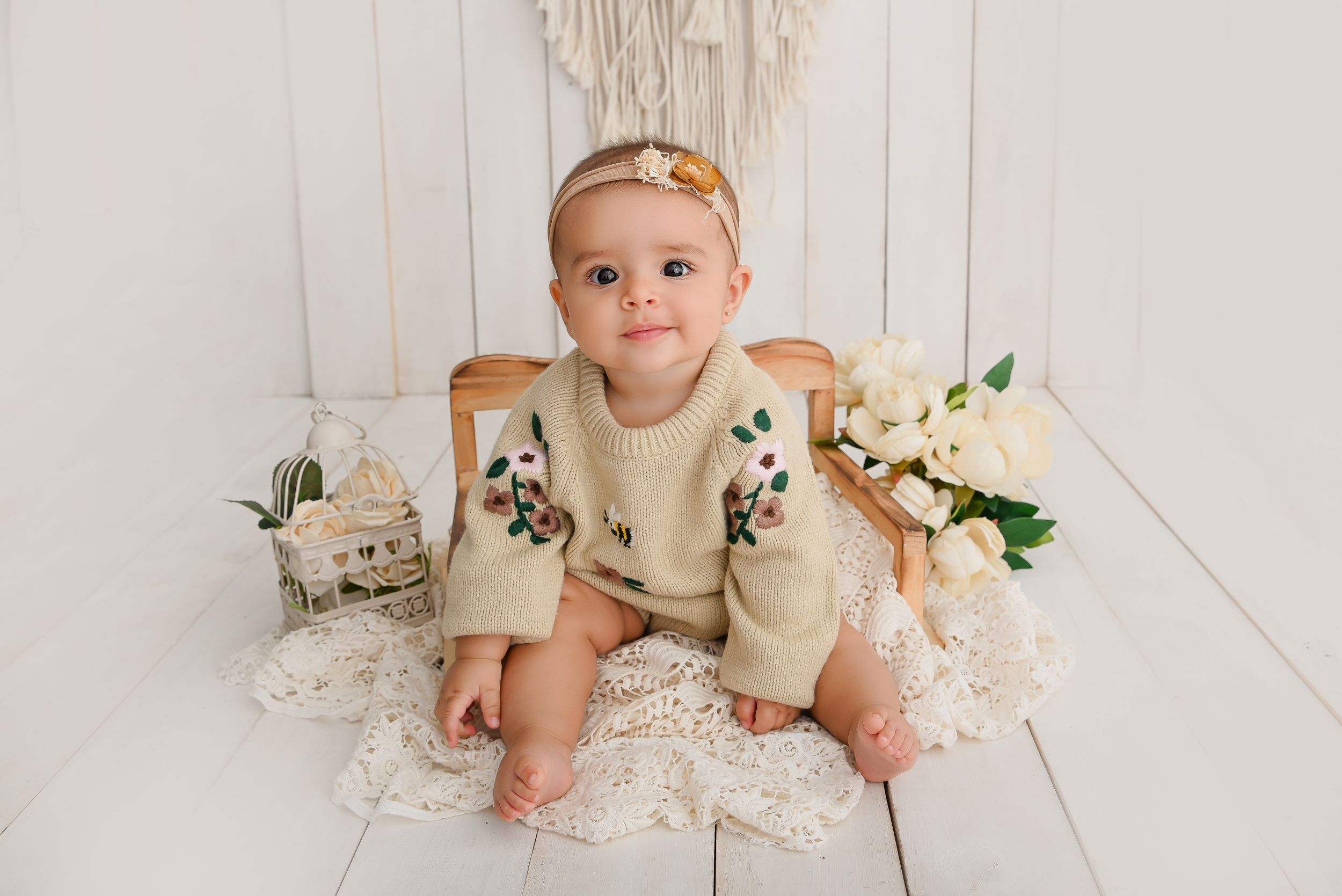 Small child wearing cream colored sweater with floral embroidery sits on white wooden floor with decorative elements.