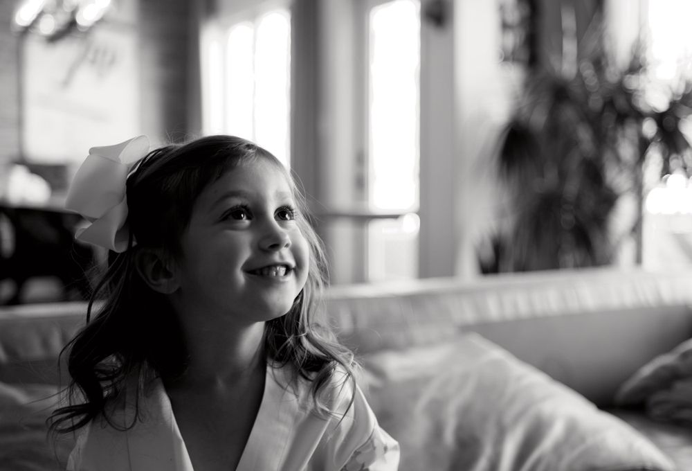 Black and white portrait of a smiling child glancing upward while sitting indoors near a window.