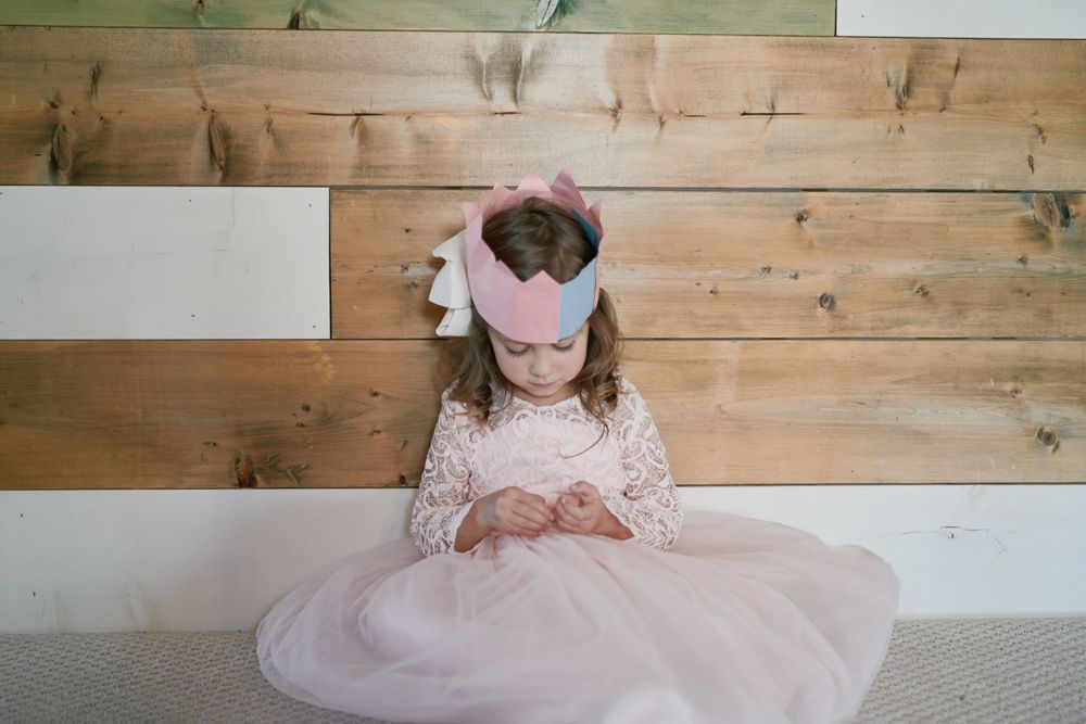 A small child wearing a tulle pink dress and bow sits on white steps against a wooden wall looking downward.