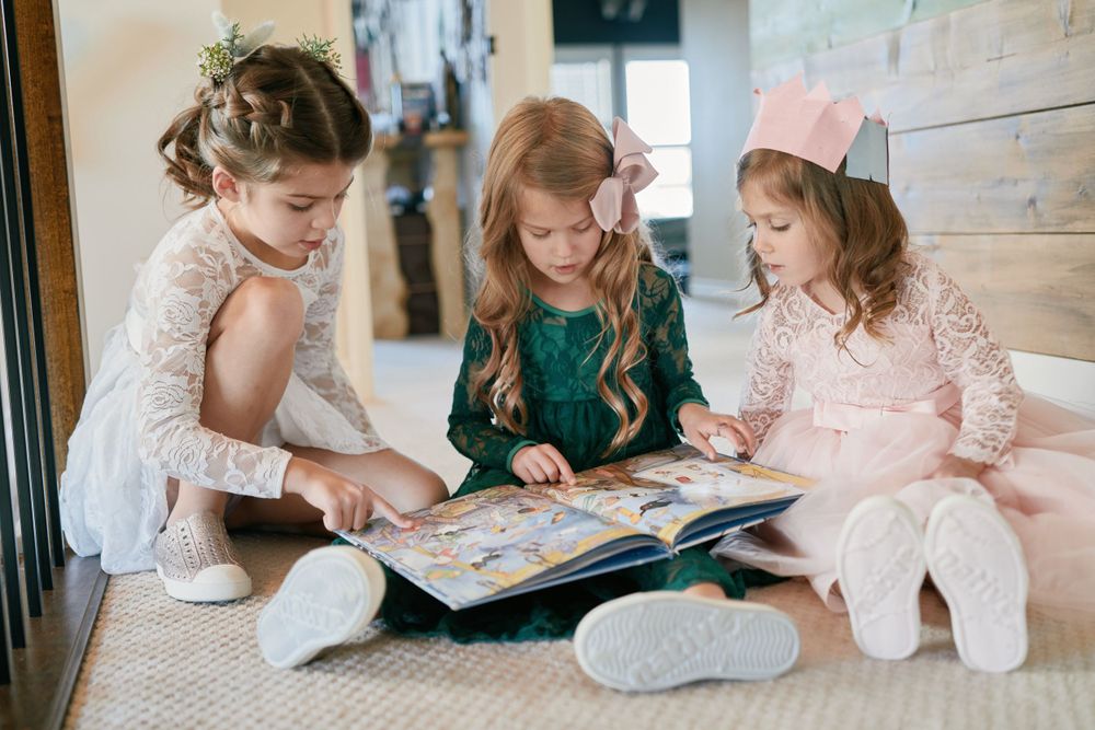 Three young children in elegant dresses sit together on the floor reading a large picture book.