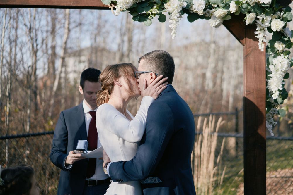 A couple shares a kiss during their outdoor wedding ceremony under a floral arch on a crisp autumn day.