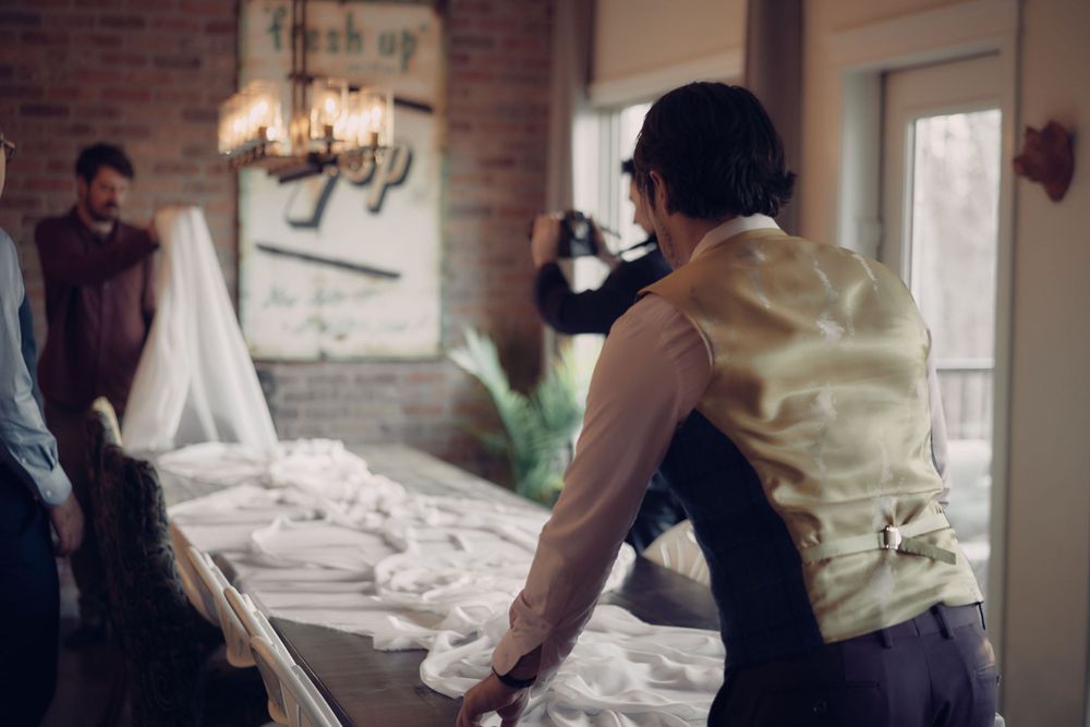 People work to prepare and set up wedding tables in a room with exposed brick walls and a hanging chandelier.