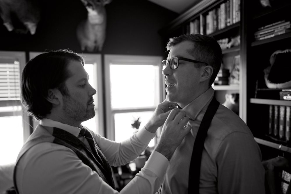 A black and white photo showing two people in formalwear adjusting a tie during wedding preparations.