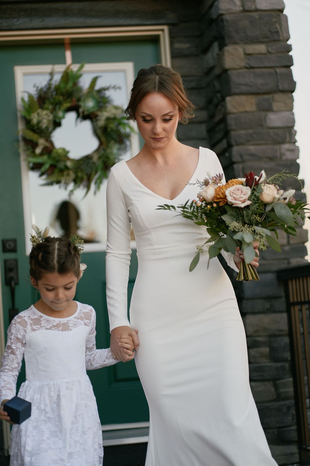 A bride in a long-sleeve white wedding dress holds a floral bouquet while standing beside a small girl in white lace.