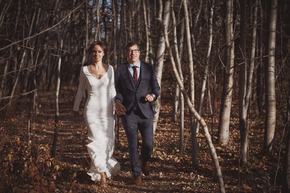A couple in wedding attire walks together through a serene autumn forest with tall birch trees in the background.